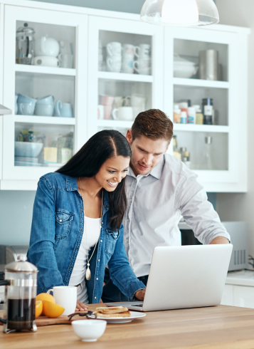 A couple stands in their kitchen reviewing information on a laptop